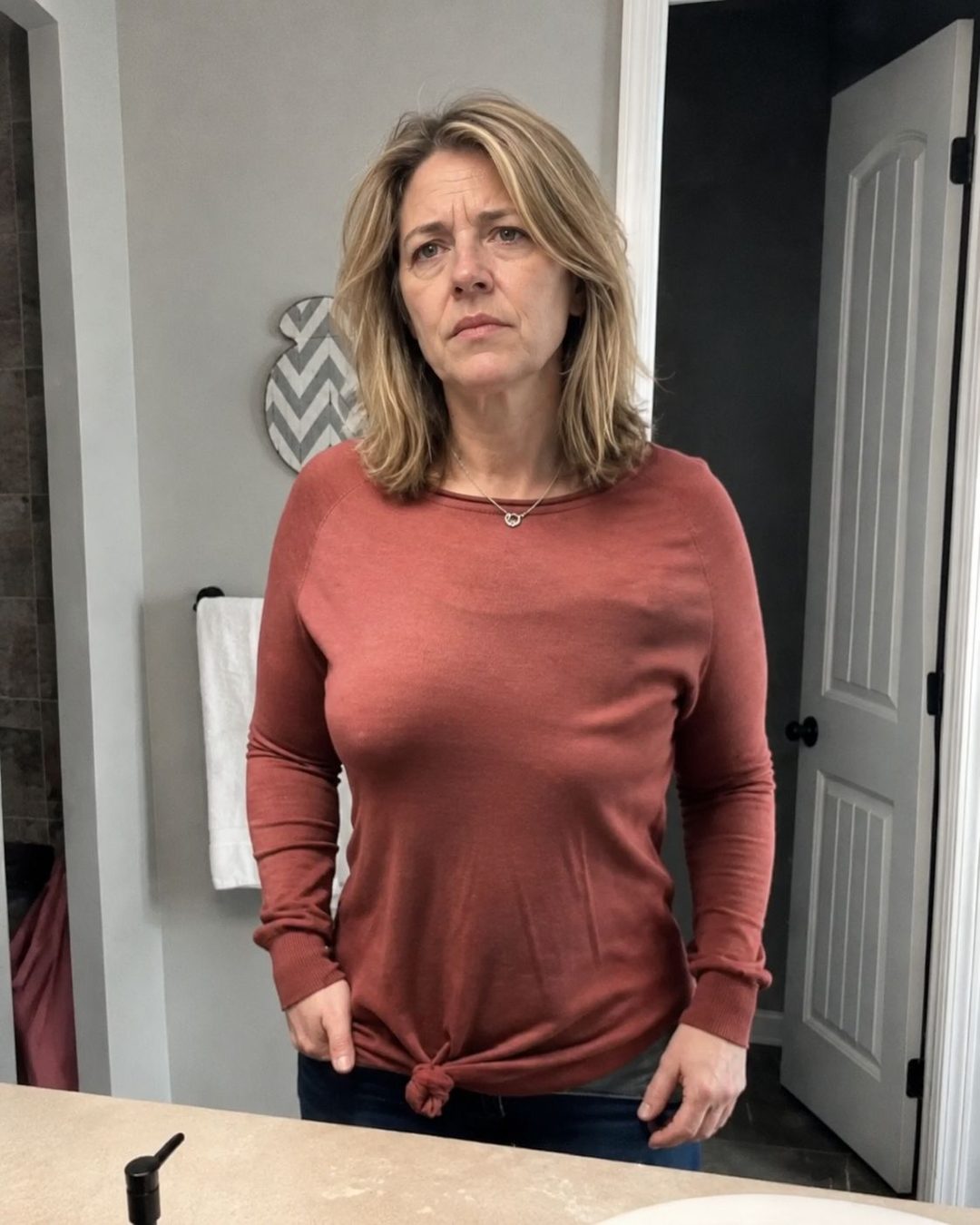 Woman in a fitted red knit top looking at herself in the bathroom mirror, unhappy with how it sits